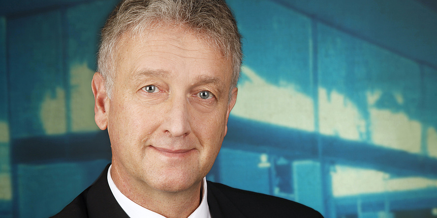 Blue background. A white haired man is smiling at the camera.