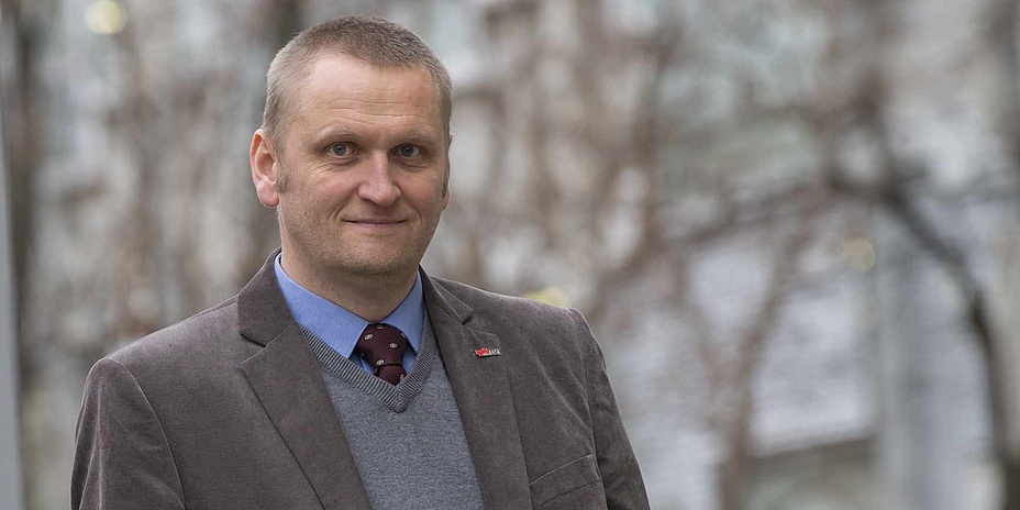 : A man with short, blond hair looks into the camera. He is wearing a blue shirt, tie and brown jacket. The photo was taken outdoors.