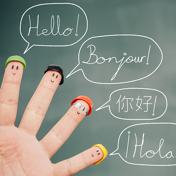 Vier Finger vor einer Tafel, auf denen Gesichter aufgemalt sind und die verschiedene Hüte tragen. Auf der Tafel steht "Hallo" in verschiedenen Sprachen. Bildquelle: lemontreeimages – Fotolia.com