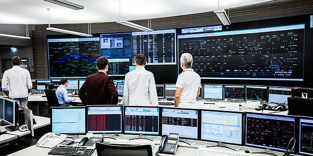 In einer Art Steuerungszentrale sind viele Bildschirme mit unterschiedlichen Zahlen und Daten zu sehen. Drei Männer blicken auf große Datenprojektionen an der Rückwand.