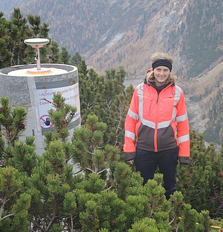 A smiling woman stands between green bushes and next to an antenna