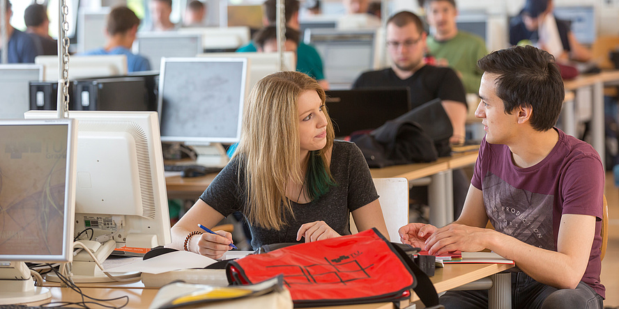 Zwei Studierende der TU Graz - ein Junge und ein Mädchen - unterhalten sihc an einem Computerarbeitsplatz in einer universitären Lernumgebung.