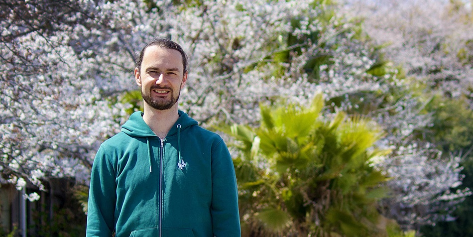 Ein Mann mit türkisem Hoody steht im Freien. Im Hintergrund sind blühende Pflanzen zu sehen. Der Mann trägt einen Bart und blickt in die Kamera. 