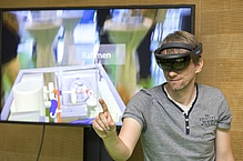 Man with virtual reality glasses and raised hand, behind him a big screen.