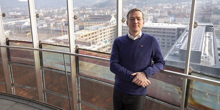 A man - Alexander Passer - stands leaning against a railing. Behind him is a glass façade that offers a view of the city. 