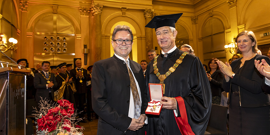 Two men smile into the camera, the man on the right wears a academic hat and holds a decoration of honor in his hand