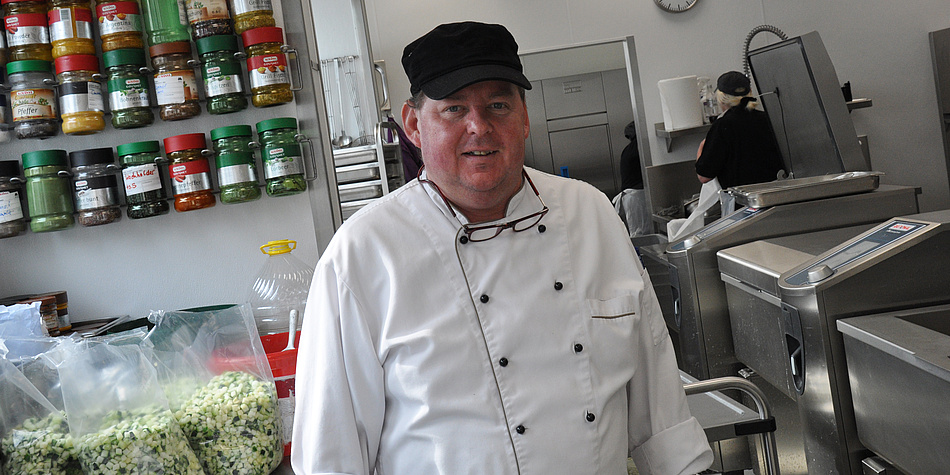 Martin Grabenhofer in white chef’s uniform and black cap in front of a shelf bearing colourful spice jars and bags of chopped vegetables in the kitchen of the Inffeldgasse canteen.