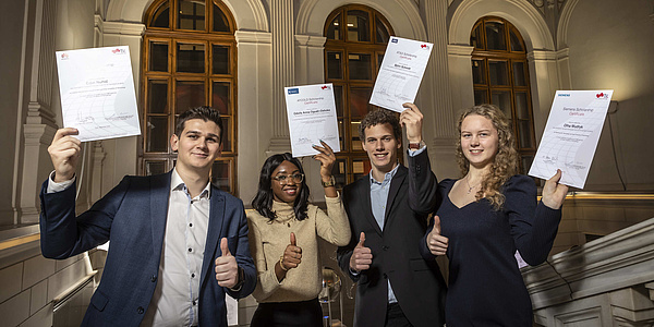 Two young women and two young men on a staircase in a historic building hold scholarship certificates up to the camera.