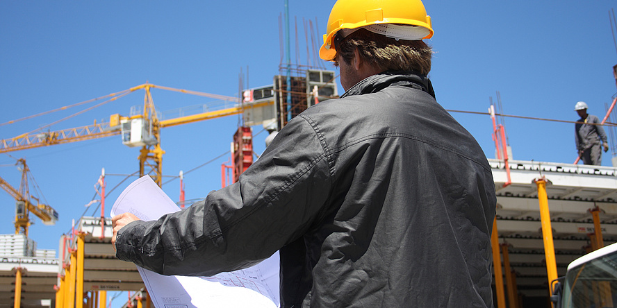 Mann mit gelbem Helm und grauer Arbeitsjacke mit dem Rücken zur Betrachterin oder Betrachter gedreht, vor einer Baustelle mit zwei Kränen schaut in einen Bauplan.