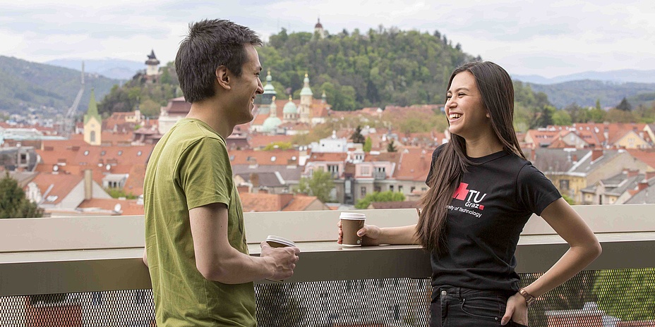 Young man and woman standing with a coffee on the railing of a terrace above the roofs of a historic old town.