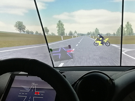 An intersection on an open country road is shown from the cockpit perspective; a yellow motorcycle is approached from the right; a display is to be found on the windscreen showing the intersection and the motorcycle as a red dot; the intersection is also shown virtually on the instrument panel behind the steering wheel.