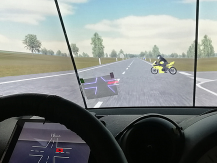 An intersection on an open country road is shown from the cockpit perspective; a yellow motorcycle is approached from the right; a display is to be found on the windscreen showing the intersection and the motorcycle as a red dot; the intersection is also shown virtually on the instrument panel behind the steering wheel.