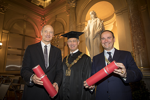 Rektor Kainz im schwarzen Talar, neben ihm Roland Busch und Robert Fischer, die jeweils eine rote Rolle in Händen halten.