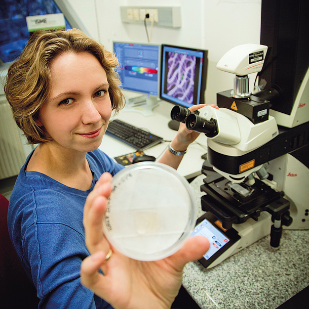 Eine junge Frau sitzt vor einem Mikroskop und präsentiert eine Petrischale.