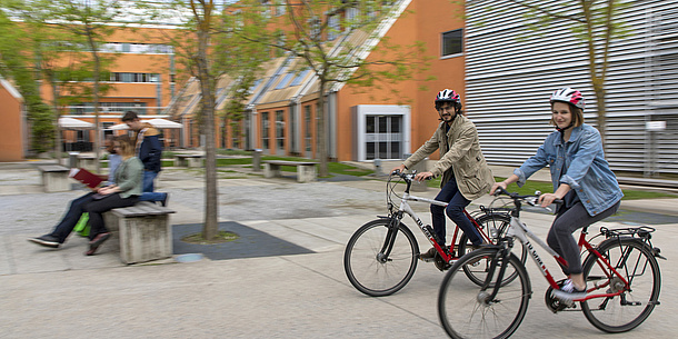 On a courtyard with some trees, two students are riding their TU Graz bicycles. In the background, students are sitting on a bench.