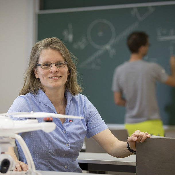A woman with a drone, behind her a young man at the blackboard.