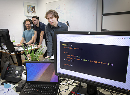 Rechts vorne ist eine Schreibtischsituation zu sehen - Am Bildschirm ist ein Computercode abgebildet, links hinten sitzen zwei Männer, ein dritter Mann steht daneben.