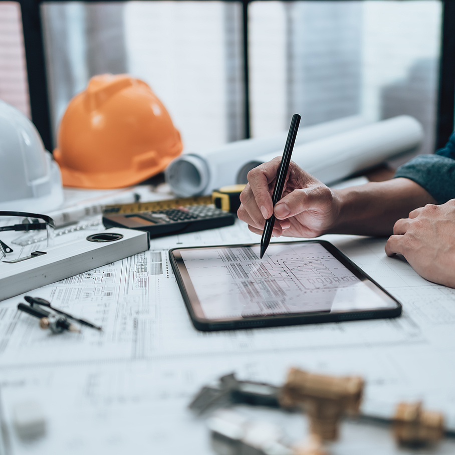 Bauhelm im Hintergrund, Hand zeichnet auf Tablet und Plan im Vordergrund