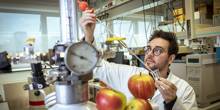Niklas Pontesegger steht in einem weißen Labormantel im Labor und hält eine Pipette. Vor ihm liegen drei Äpfel.