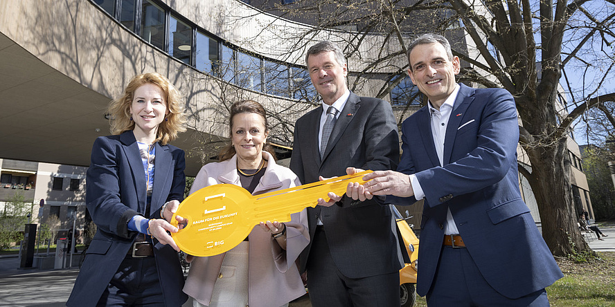 Four people are smiling at the camera: two women on the left and two men on the right. They are holding a yellow key between them.