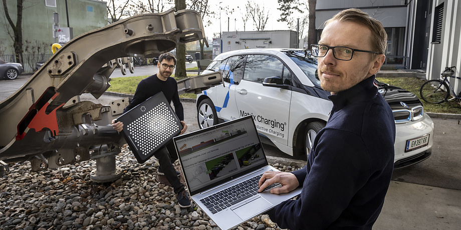 Two men stand in front of a car and a construction part of a railway. The man on the left holds a board with metallic circles, the man on the right holds a computer