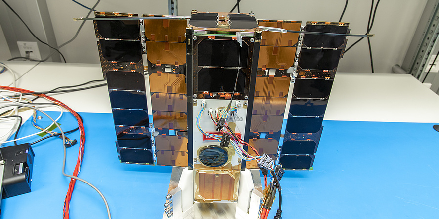 A small satellite with a few cables attached stands on a table with a blue pad. 