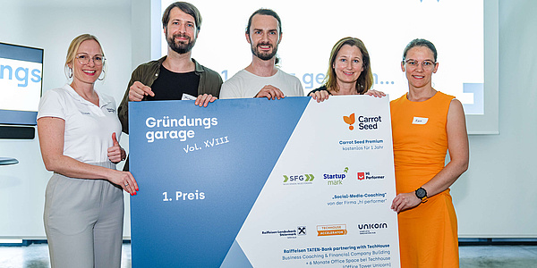 Zwei Männer und drei Frauen halten ein Plakat mit Unternehmenslogos und der Aufschrift „Gründungsgarage 1. Preis"