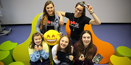 Vier Mädchen im Schulalter von 10 bis 14 Jahren und eine Frau mit Brille präsentieren jeweils eine Computerplatine, ein Tablet und einen drei Zentimeter großen kugelförmigen Roboter.
