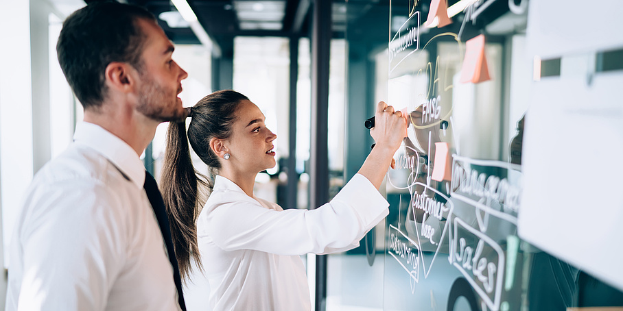 Side view of female drawing scheme of business strategy on glass board while male coworker observing process at modern workplace