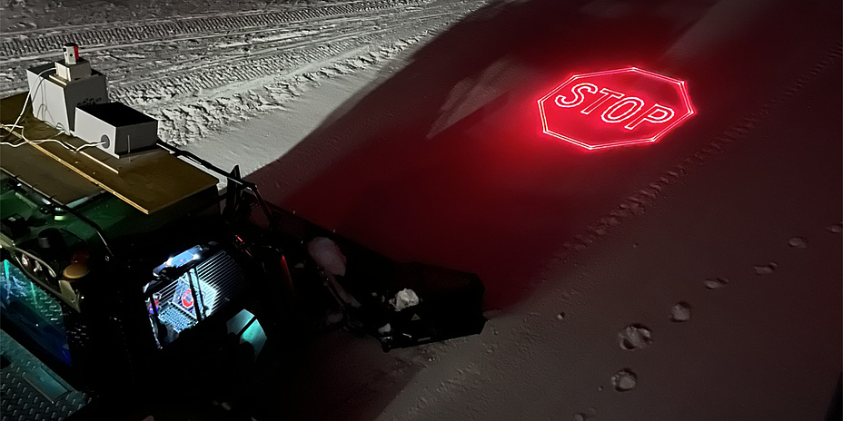 Eine Pistenraupe fährt von links in das Bild. Vor ihr ist in Rot das Wort "Stop" in den Schnee projiziert.