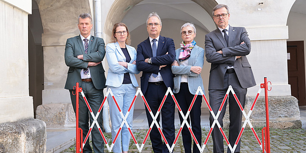 Five people are standing with their arms crossed behind a waist-high barrier. There are three men and two women.