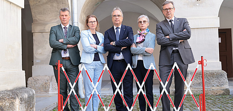 Fünf personen stehen mit verschränkten Armen hinter einer hüfthohen Absperrung. Es sind drei Männer und zwei Frauen.