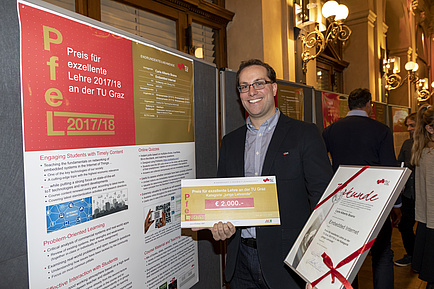 A man is holding a tribute in his right and left hand. To the left of him is a poster wall showing a poster in A1 format.