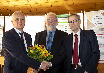 Drei Männer in Anzügen schütteln einander die Hände, der linke bekommt einen Blumenstrauß überreicht