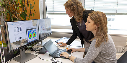 Zwei Frauen an einem Arbeitsplatz mit Laptop und zwei Bildschirmen.