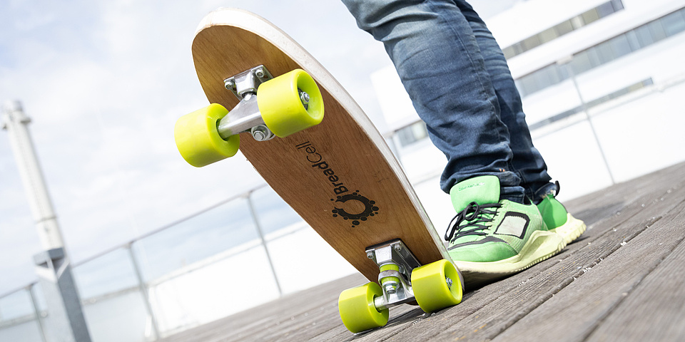 Ein Skateboard mit grünen Rädern, dessen Ende jemand mit einem Fuß hinunter drückt, sodass es vorne nach oben steht. Unten auf dem Skateboard ist das Wort "BreadCell" lesbar.