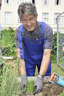 Mann mit dunklen Haaren und blauer Latzhose trägt graue Gartenhandschuhe und gartelt. 