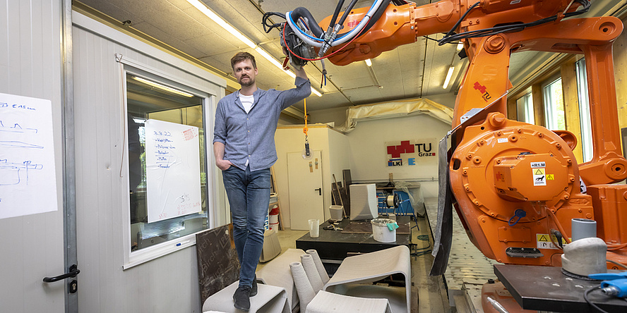 Ein Mann steht auf einem Beton-Objekt. Seine Hand liegt auf dem Kopf eines großen, organgen Roboterarns.
