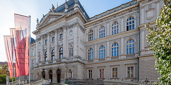 Three flags fly from flagpoles in front of a three-storey historic building.