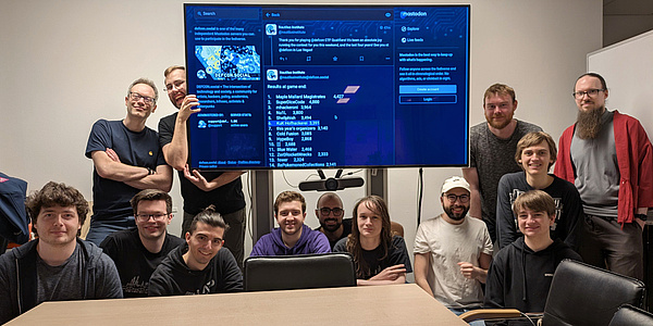 Hinter einem Tisch hocken bzw. stehen 13 Personen um einen großen Flachbildschirm herum und schauen in die Kamera.