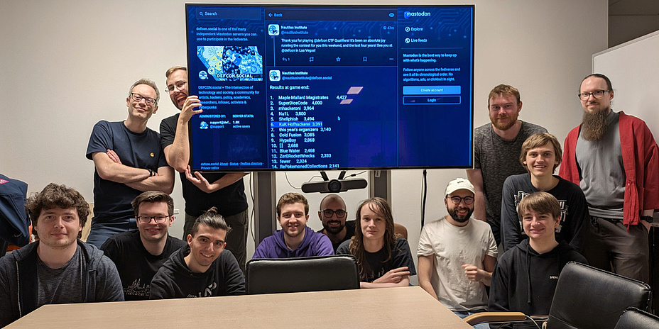 Hinter einem Tisch hocken bzw. stehen 13 Personen um einen großen Flachbildschirm herum und schauen in die Kamera.