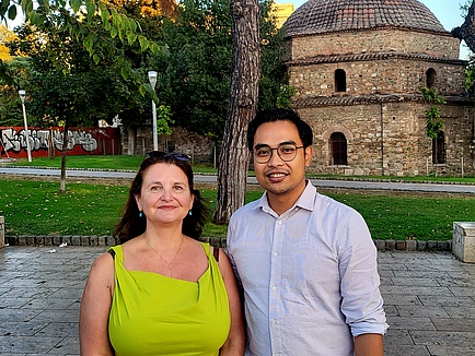 A woman and a man stand in front of an old building and smile into the camera