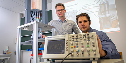 A man in a chequered shirt is standing next to a man in a grey shirt. In front of her stands a device with many vertical tubes.