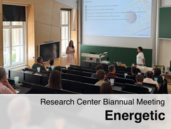 A room full of people listening to a lecture during the Research Center Biannual Meeting "Energetic". Two women are standing at the front of the room, one of whom is giving a presentation. The meeting's agenda is displayed on a screen.