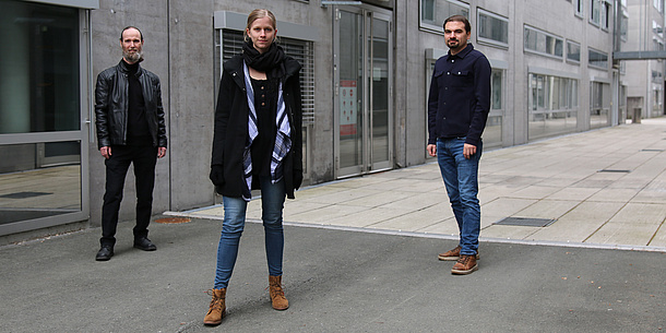 Eine Frau und zwei Männer stehen vor einem grauen Gebäude.