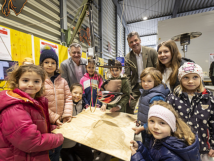 Zahlreiche Kinder und zwei Männer stehen um Holzplatte, über der ein künstlicher Kopf mit Fahrradhelm hängt.