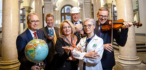 Zwei Frauen und vier Männer stehen als Gruppe zusammen. Eine Person hält einen Globus, eine eine Geige, eine hat einen Bauhelm am Kopf und ein Stück Gestein in der Hand, eine hält ein Computerbauteil, eine trägt einen Arztmantel und ein Stethoskop und eine hält eine kleine Drohne in der Hand.