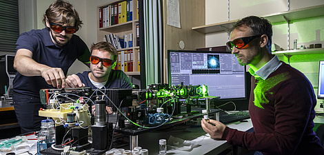Drei Männer tragen Schutzbrillen und hantieren an einem Laborgerät, durch dessen Glasröhrchen ein Laserstrahl hindurchleuchtet.