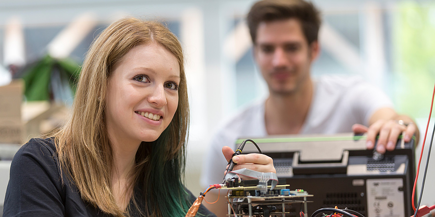 A young woman with long blond hair in the foreground and a dark-haired young man in the background work on a sensor