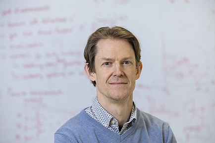 A man standing in front of a blackboard covered in writing looks into the camera.
